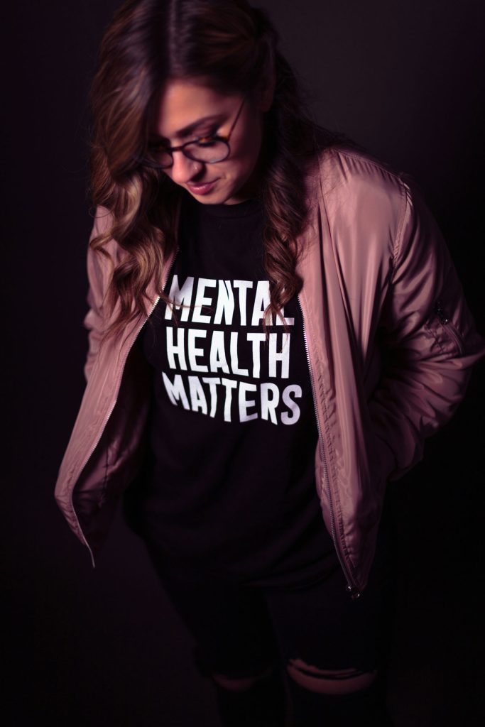 A young woman with wavy brown hair and glasses looks down with a gentle expression, wearing a black T-shirt that boldly states 'Mental Health Matters.' She is dressed in a soft pink bomber jacket, and the dim lighting gives the image an introspective and powerful mood. This image represents the importance of prioritizing survivors' mental health, emphasizing awareness, healing, and self-care after trauma.