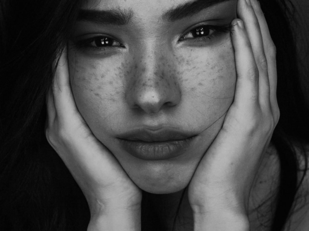 "A black-and-white close-up of a young woman with freckles, her face resting in her hands. Her eyes appear glossy and distant, reflecting deep sadness and emotional exhaustion. The lack of expression and hollow gaze powerfully depict the aftermath of emotional abuse, capturing the silent suffering and inner turmoil of someone struggling with complex PTSD."