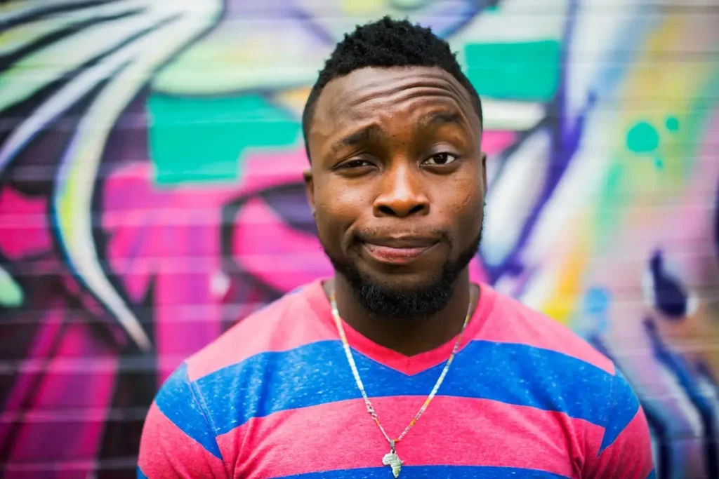 A man with short black hair and a well-groomed beard stands in front of a vibrant graffiti wall, wearing a pink and blue striped shirt. He raises one eyebrow and smirks slightly, conveying skepticism or confusion. His facial expression reflects uncertainty, often purposely done in narcissistic relationships when someone is setting a boundary to make them feel crazy and second-guess themselves.