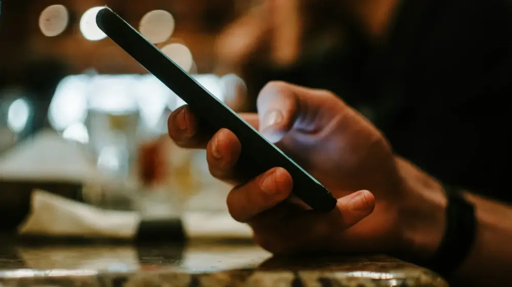 A close-up of a man's hand holding a smartphone, their thumb hovering over the screen in a dimly lit setting. The blurred background suggests a public place, possibly a café or bar. This image represents the psychological manipulation tactic known as hoovering, where an abuser reaches out through messages or social media to lure a past victim back into a toxic cycle of control and emotional abuse.