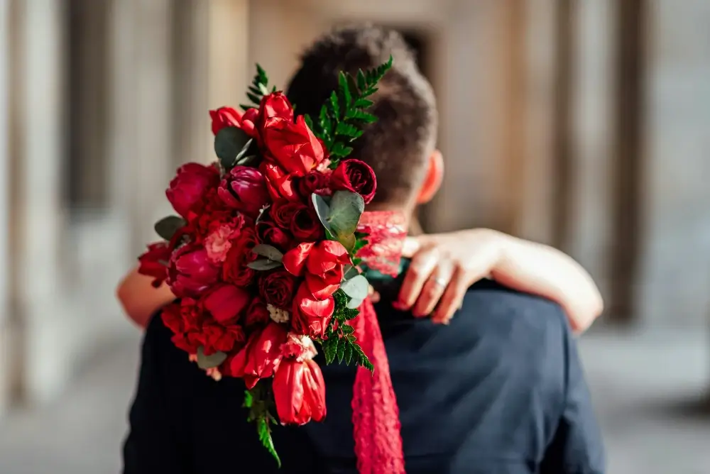 Woman embraces a man dressed in a dark suit, her hands wrapped around his neck while holding a bouquet of vibrant red roses and peonies. The deep red tones of the flowers symbolize passion and intensity, often associated with love bombing—a manipulative tactic where excessive affection, gifts, and grand gestures are used to gain control in a toxic relationship.