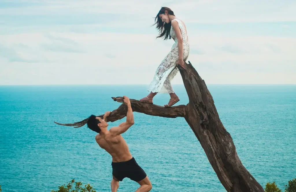 A shirtless, muscular man struggles to hold onto a precarious tree branch over a cliffside, while a confident woman in a white lace dress stands above him, looking down with an air of dominance. The vast ocean stretches behind them, symbolizing freedom and detachment. This visually striking scene metaphorically represents a man losing control over a woman who is reclaiming her power—an image that perfectly encapsulates how to piss off a narcissist by refusing to be controlled.