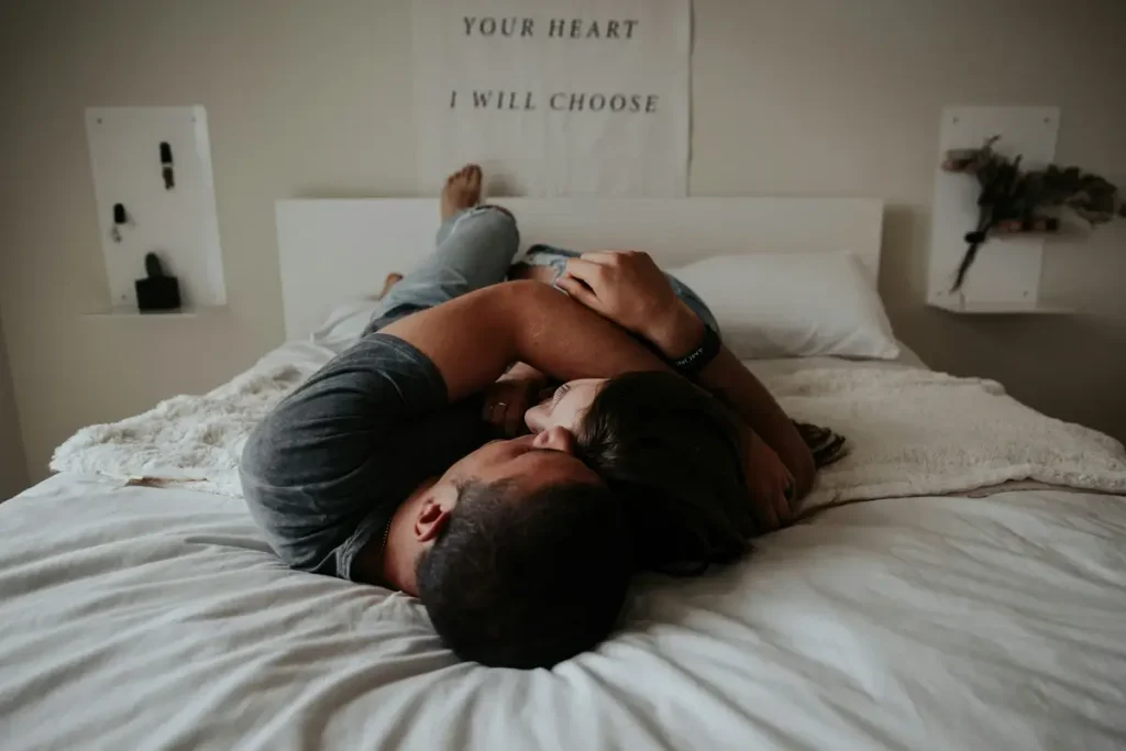 A couple lies intertwined on a cozy bed, embracing passionately in an intimate moment. The man, wearing a dark t-shirt, holds the woman close as they share a deep connection. The soft lighting and romantic atmosphere contrast with the hidden emotional toll of sex with a narcissist, where physical closeness can mask emotional manipulation.