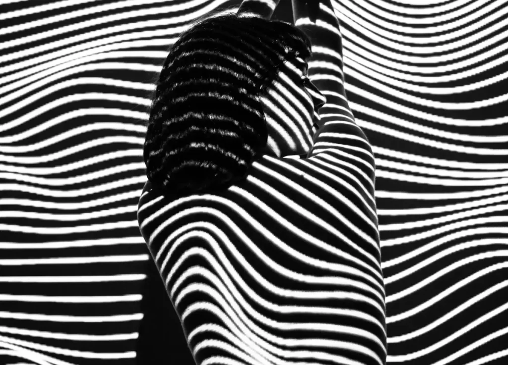 A woman stands with her arms raised, her body and face covered in high-contrast black and white striped shadows, creating a fragmented visual effect. This striking image represents narcissist splitting, black and white thinking, psychological concepts where individuals perceive others in extremes.