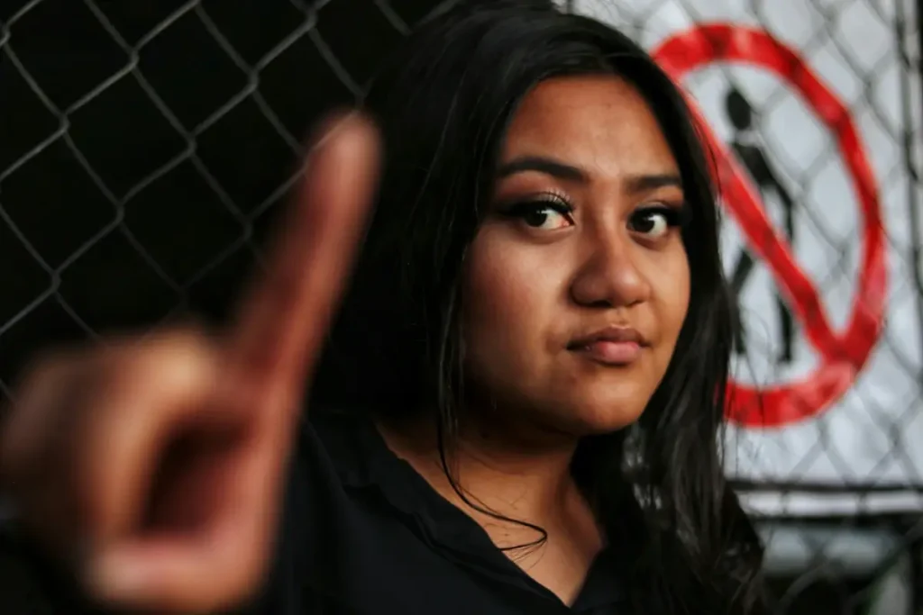 A serious-looking woman with long dark hair stands in front of a chain-link fence with a 'no entry' sign, holding up her finger in a firm 'stop' gesture. Her facial expression conveys determination and assertiveness, symbolizing the act of setting boundaries. This image represents the concept of enforcing personal limits, a crucial aspect of learning how to set boundaries with a narcissist.