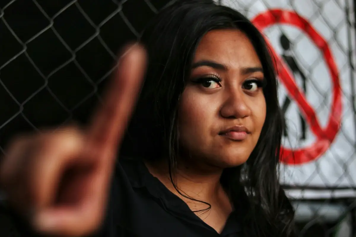 A serious-looking woman with long dark hair stands in front of a chain-link fence with a 'no entry' sign, holding up her finger in a firm 'stop' gesture. Her facial expression conveys determination and assertiveness, symbolizing the act of setting boundaries. This image represents the concept of enforcing personal limits, a crucial aspect of learning how to set boundaries with a narcissist.