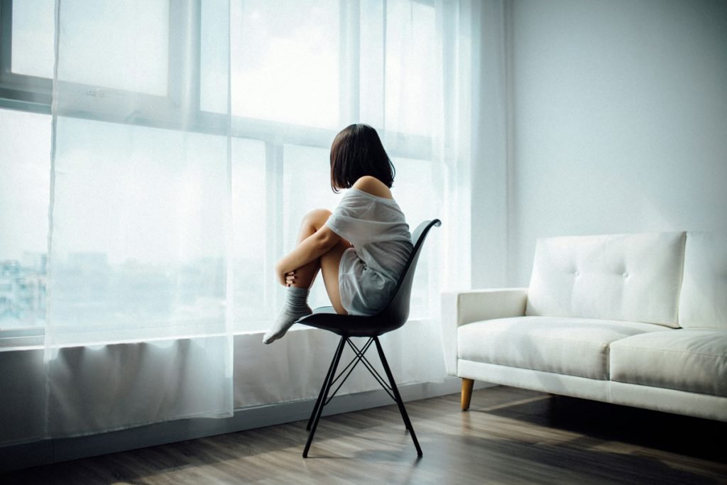 A young woman sits curled up on a black chair near a large window, gazing outside with a distant expression. She wears an oversized off-the-shoulder shirt and socks, conveying a sense of isolation and emotional distress. The minimalistic, neutral-toned living space enhances the feeling of loneliness. This image captures the deep emotional exhaustion and confusion often felt by someone who has been hoovered by a narcissist, struggling to regain their sense of self after manipulation.