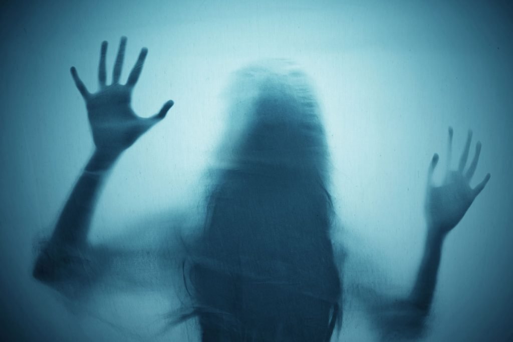 A blurred silhouette of a woman with long hair presses her hands against a fogged or frosted glass surface, her fingers spread wide in a desperate gesture. The eerie blue tint and distorted outline evoke feelings of fear, entrapment, and isolation. This powerful image symbolizes the emotional turmoil of a trapped abuse survivor, struggling to break free from the cycle of manipulation and control.